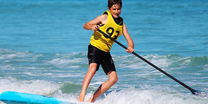 Un jeune surfeur sur sa planche de paddle glisse sur les vagues turquoise d'une plage ensoleillée, illustrant l'énergie et l'esprit des sports nautiques d'été.