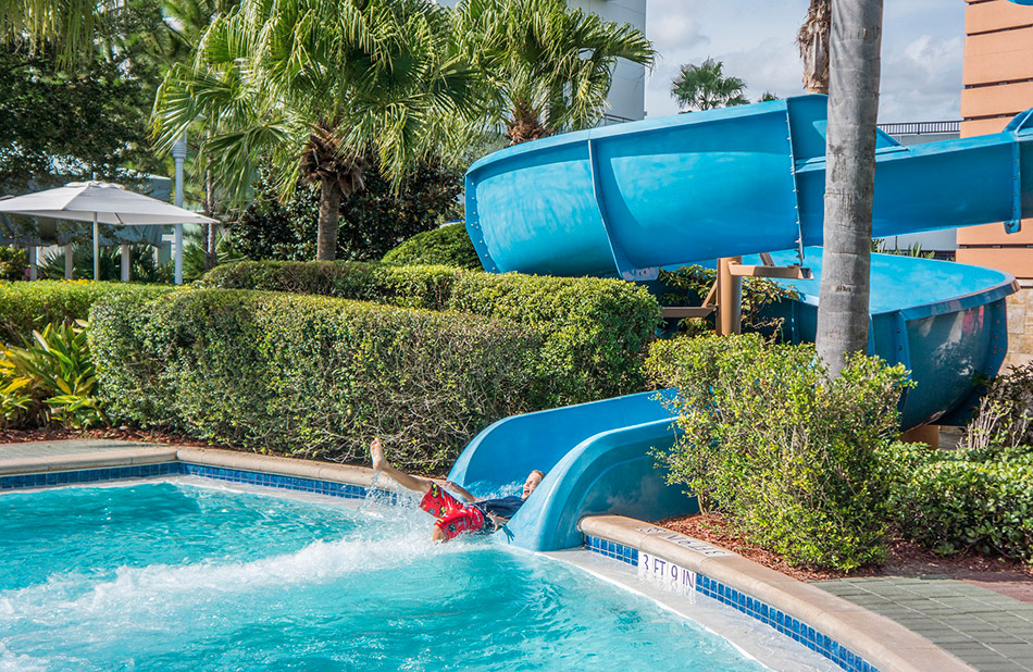 Enfant glissant joyeusement sur un toboggan bleu dans un parc aquatique ensoleillé, entouré de palmiers, créant une scène estivale parfaite pour des souvenirs en famille.