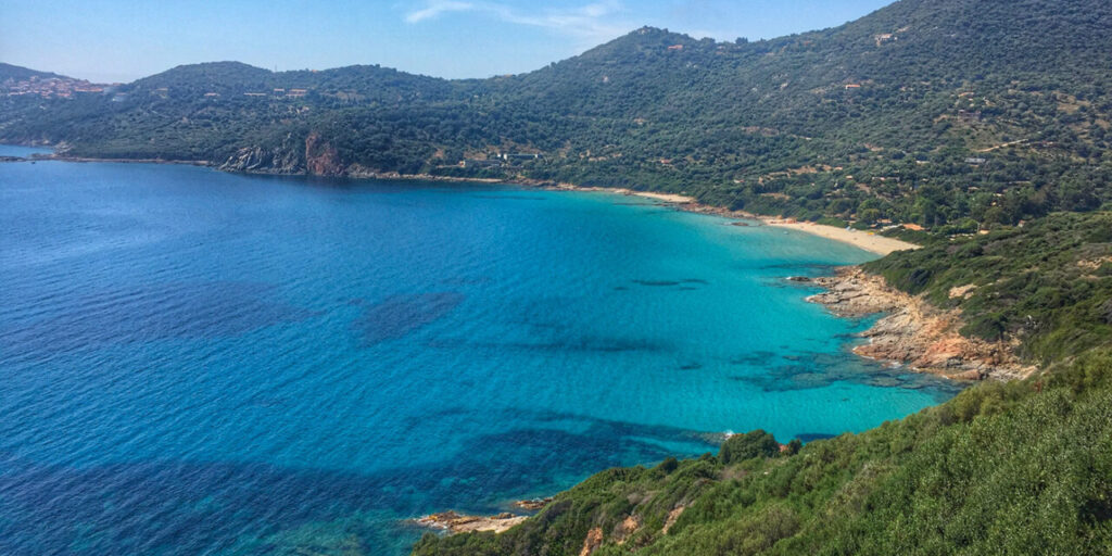 Image d'une plage de sable fin en Corse, bordée par des eaux cristallines et surmontée de collines verdoyantes, représentant la beauté sereine de la Méditerranée.