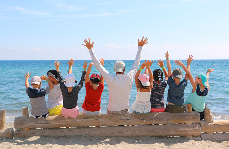 Des enfants joyeux, bras levés, jouent avec leurs aînés sur une plage ensoleillée, illustrant le bonheur estival en famille.
