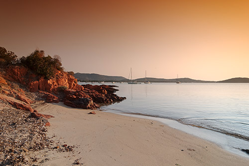 coucher-de-soleil-sur-une-plage-tranquille Coucher de soleil enchanteur sur une plage calme avec voiliers à l'horizon, reflet doré sur l'eau.