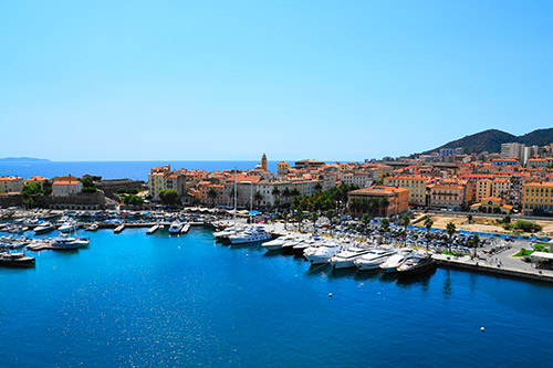 Vue panoramique du port marin de Corse avec bateaux sur mer, ciel bleu dégagé et architecture colorée en arrière-plan.