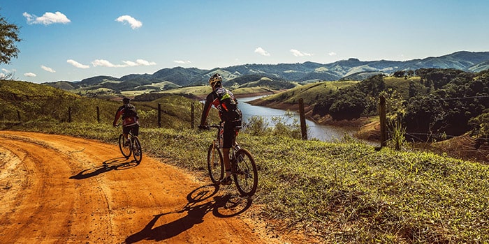 Deux cyclistes s'aventurent sur un sentier pittoresque entouré de montagnes verdoyantes, profitant d'une journée ensoleillée et d'une vue panoramique sur un lac scintillant.