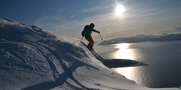 Skieur dévalant une montagne enneigée avec une vue imprenable sur la mer scintillante sous le soleil, incarnant l'esprit d'aventure et l'amour de la nature.