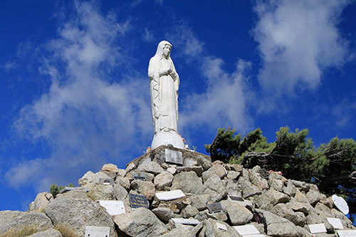 Statue de la Vierge sur une montagne, offrant une vue panoramique sous un ciel bleu méditerranéen. Symbole de foi, elle évoque sérénité et spiritualité.