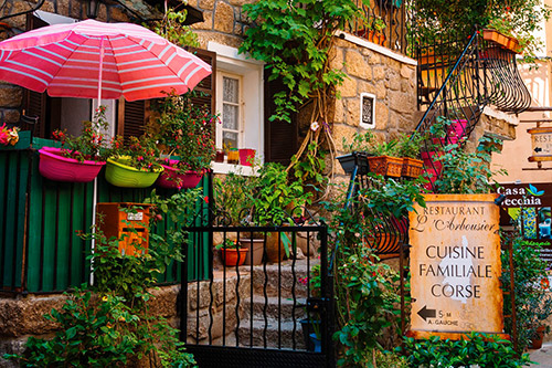 Cette image représente une façade de restaurant traditionnel en Corse, ornée de fleurs colorées et d'éléments pittoresques, créant une atmosphère chaleureuse et accueillante.