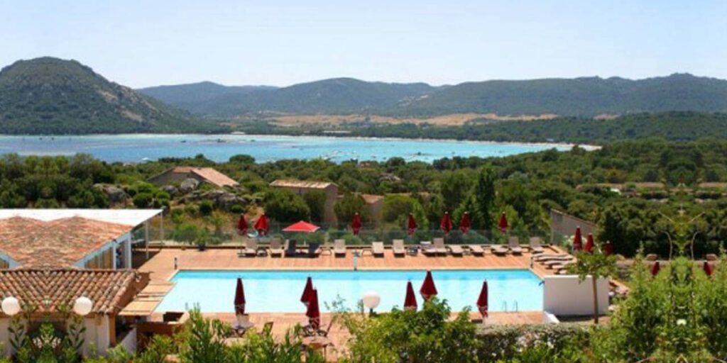 Vue imprenable d'une piscine d'hôtel avec parasols rouges, surplombant la mer et les montagnes verdoyantes, idéale pour des vacances relaxantes.