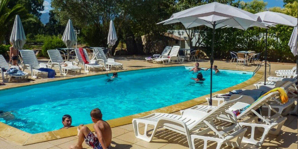 Enfants jouant dans une piscine sous le soleil Des enfants s'amusent dans une piscine éclatante de soleil, entourés de chaises longues et de parasols offrant détente et ombre.