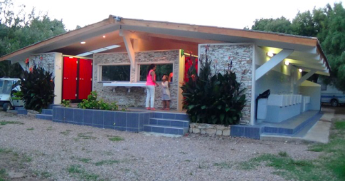 Bâtiment moderne de camping au crépuscule Bâtiment de camping moderne avec murs en pierre et portes rouges illuminées au crépuscule, entouré de plantes luxuriantes, créant une atmosphère chaleureuse et conviviale pour les campeurs.