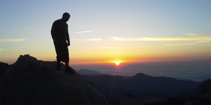 Cette image capture un homme en silhouette sur une montagne, contemplant un spectaculaire coucher de soleil aux teintes vives d'orange et de rose, symbolisant la connexion avec la nature.