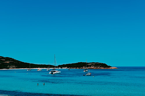 Cette image illustre une magnifique plage méditerranéenne avec des eaux turquoise sous un ciel sans nuages, des voiliers paisibles et des baigneurs profitant d'une journée ensoleillée.