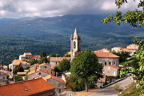 Cette image montre le village de Zonza en Corse, capturant ses maisons au toit de tuiles orangées et son imposant clocher sous un ciel nuageux, entouré par les montagnes verdoyantes.